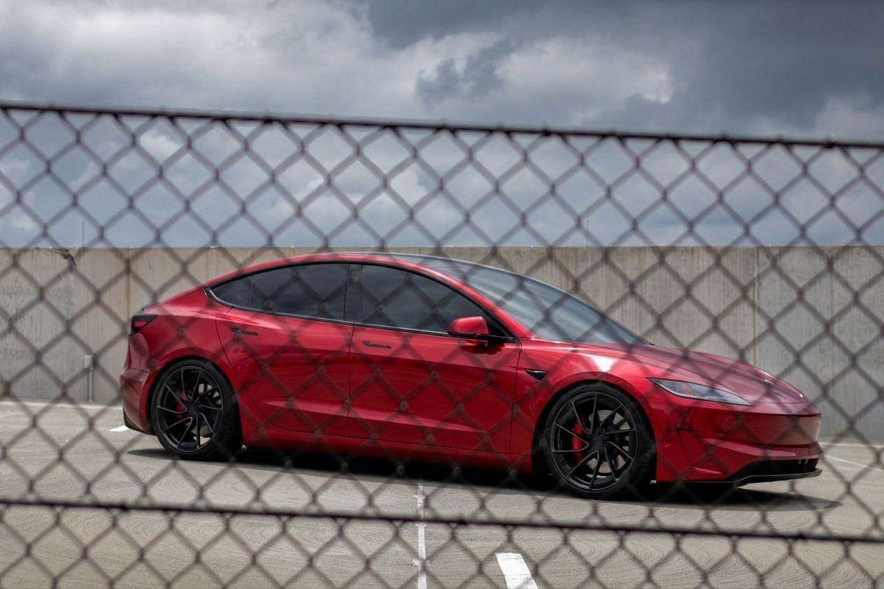 Red Tesla Model 3 parked behind a chain-link fence under storm clouds